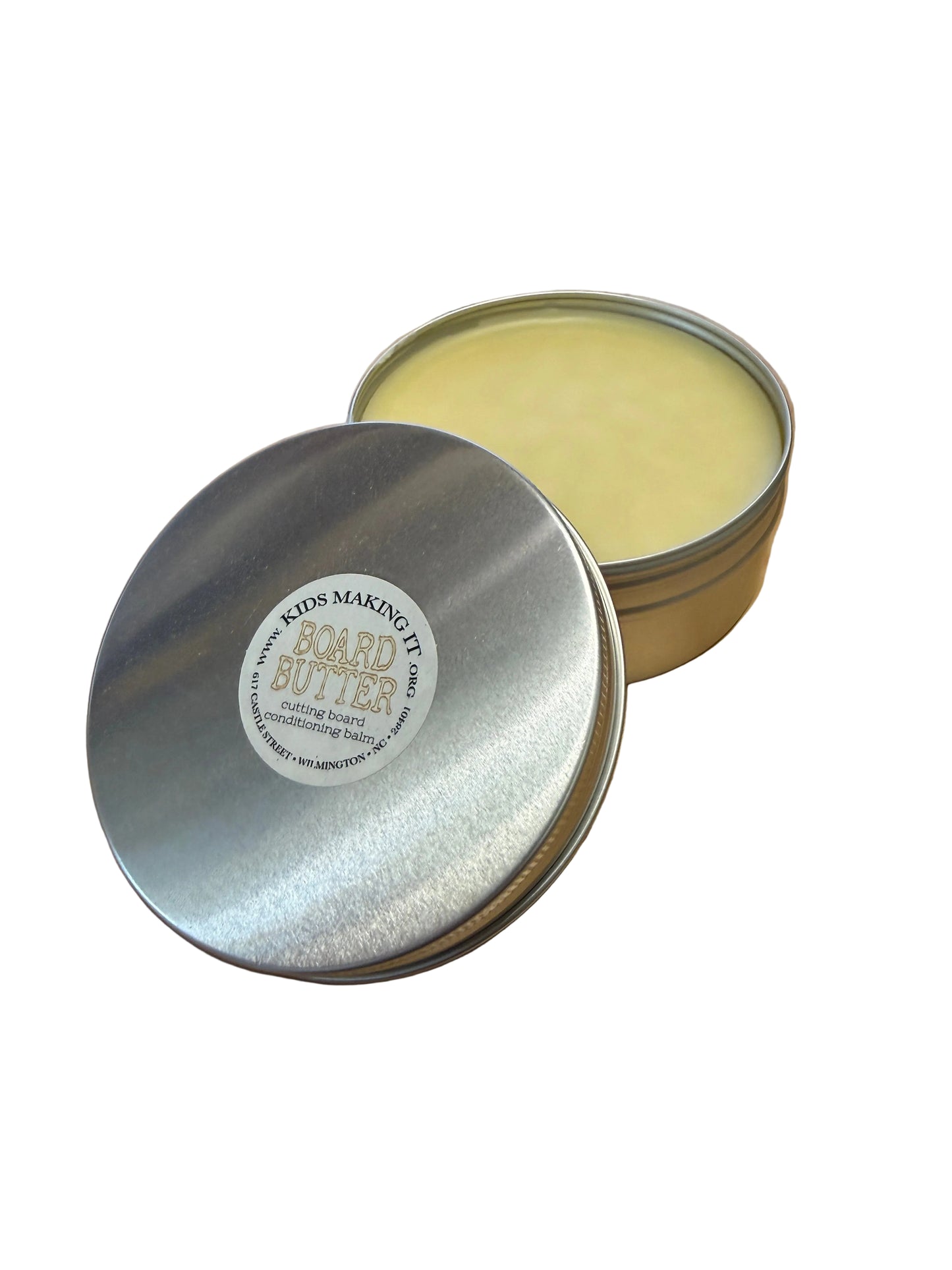 A round silver metal tin of beeswax with its lid off, showing pale yellow wax inside, placed on a wooden desk next to a laptop and mouse. The lid has a circular label reading 'BEEBUTTS BEES WAXING IT since 2011'.